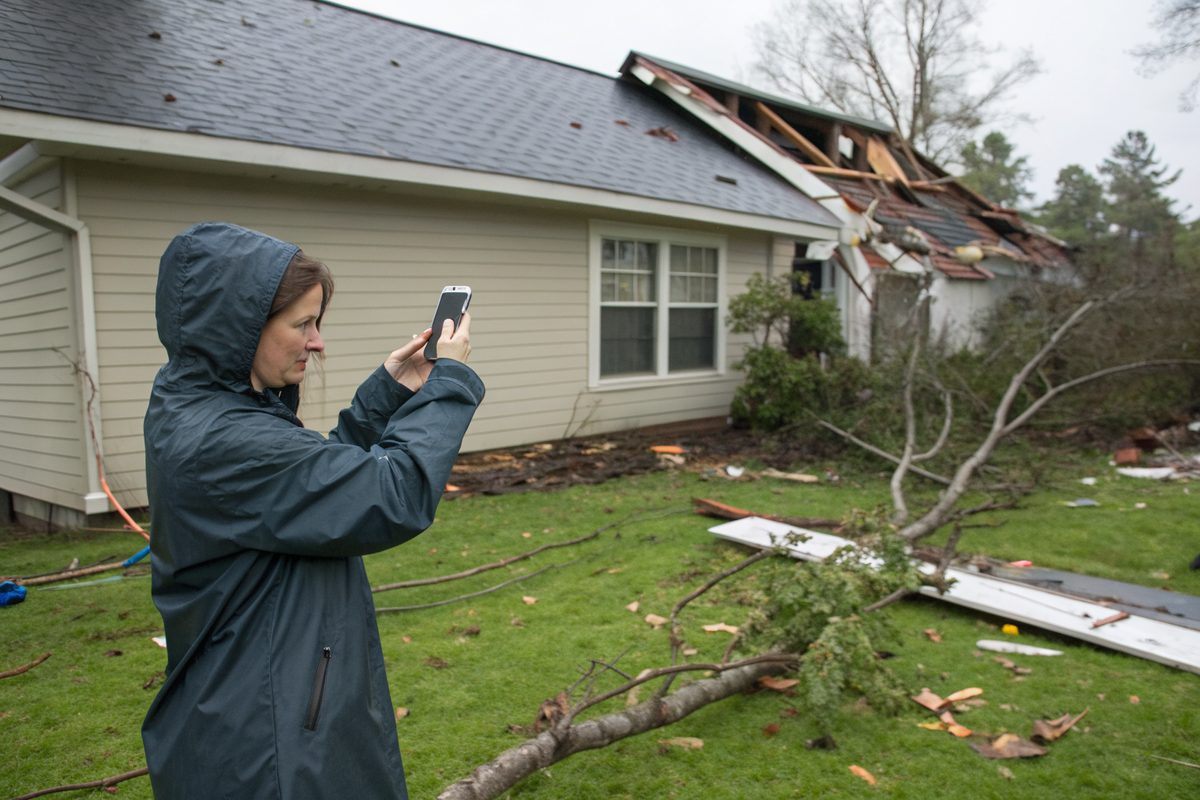 A homeowner in a rain jacket photographing wind damage to her roof, with branches on the lawn